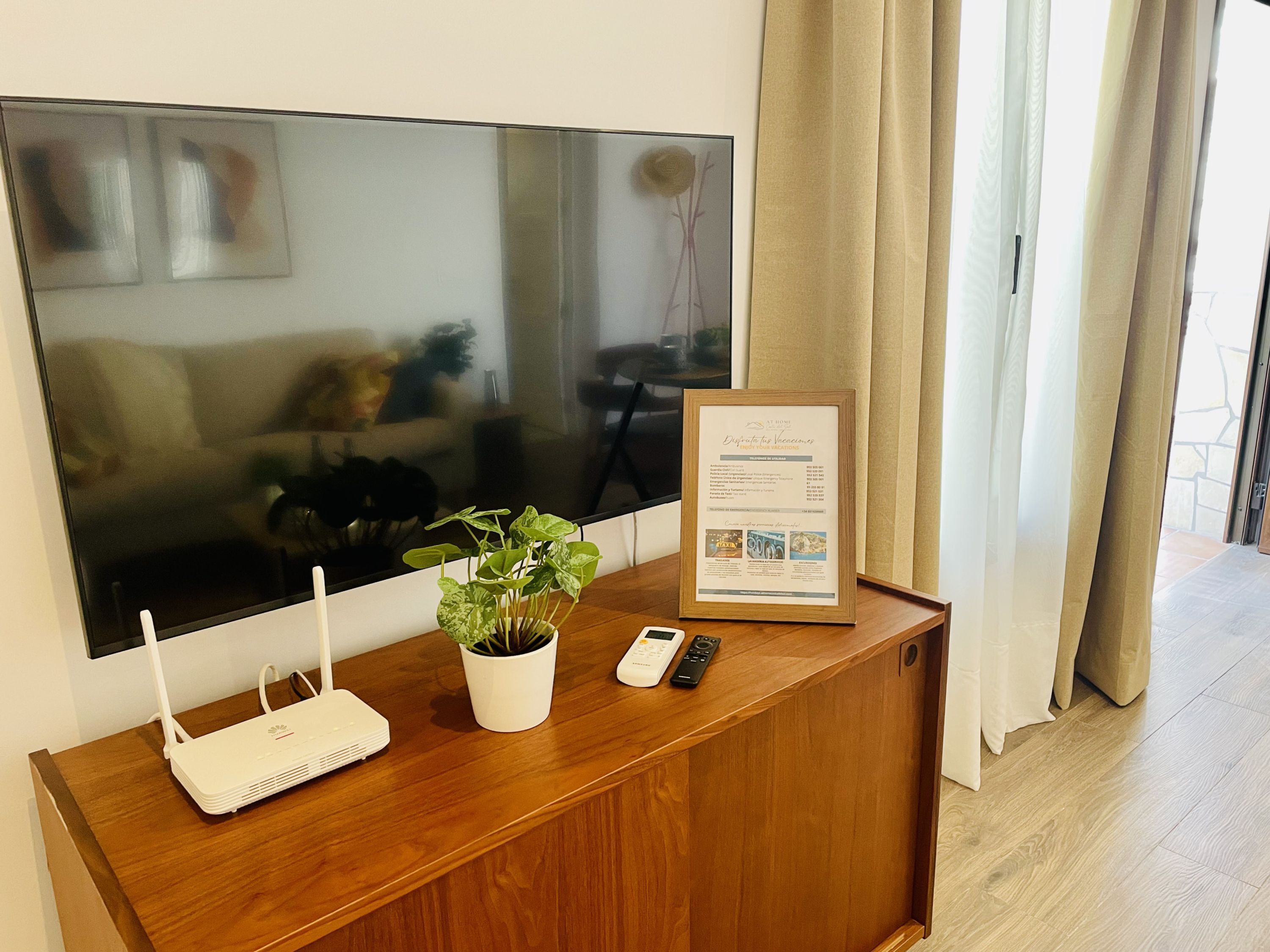 Un escritorio de madera con un router, una planta en maceta, un documento enmarcado y un control remoto.
