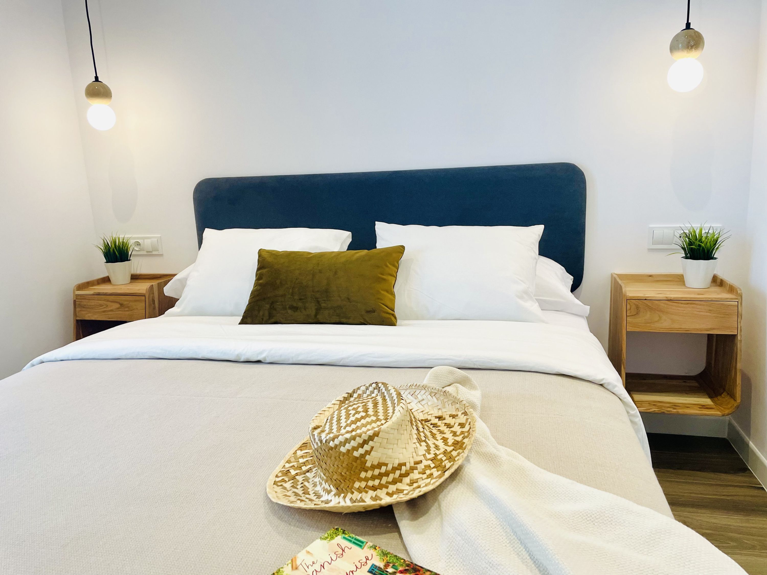 A neatly made bed with a blue headboard, white bedding, and a woven hat on the bed. There are two bedside tables with small potted plants.