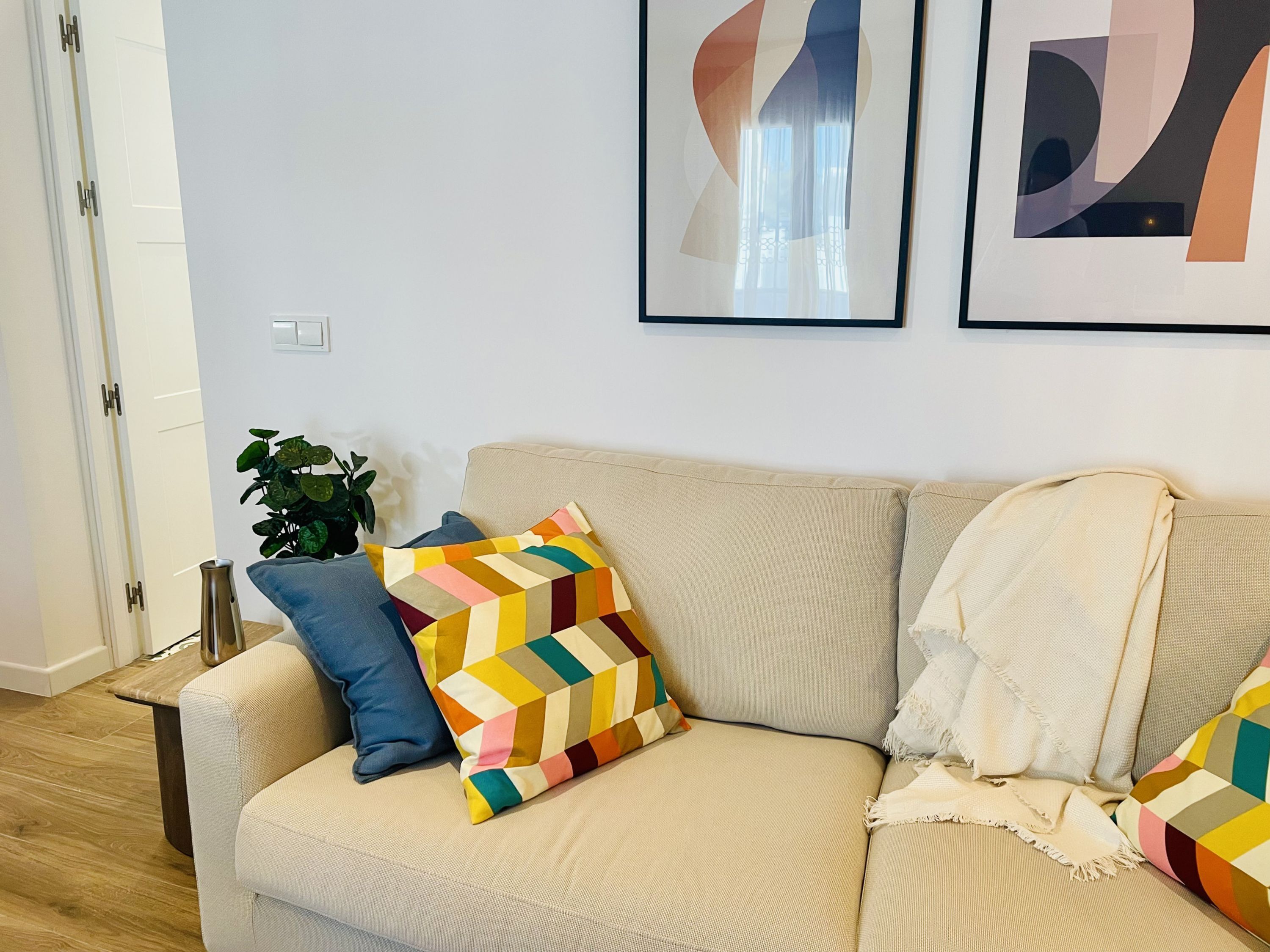 A living room with a beige sofa, colorful geometric pillows, a white throw blanket, and abstract wall art.