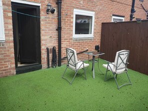 Eine Terrasse mit Kunstgr Rasen, zwei Metallstühlen, einem kleinen Tisch und einer Ziegelwand mit Fenster und Tür.