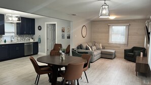 Una cucina moderna con pensili scuri, un'area pranzo con tavolo e sedie rotonde, un soggiorno con divano componibile e un lampadario a soffitto.