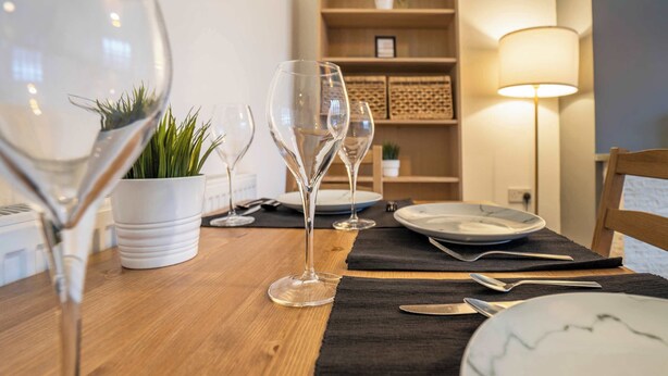 Ein Esstisch mit Weingläsern, Tellern und Besteck, einer Topfpflanze und einer Lampe in einem Raum mit Holzboden und Regalen im Hintergrund.