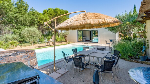 Un espace aménagé au bord de la piscine, avec une table et des chaises, un parasol en chaume et un bâtiment doté d’une porte en verre.