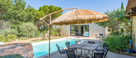 Un área junto a la piscina con mesa y sillas, una sombrilla de paja y un edificio con puerta de cristal.