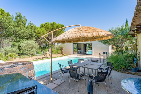 A poolside area with a table and chairs, a thatched umbrella, and a building with a glass door. - Charming Provence Villa with Pool, Wi-Fi, and Air Conditioning (Rochegude)