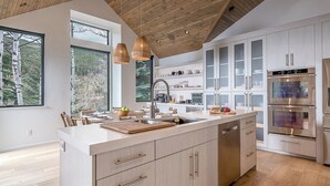 A modern kitchen with wooden cabinetry, a central island, stainless steel appliances, and large windows offering a view of trees. - Sleek, Light-Filled Alpine Escape | Hot Tub, Wine Room | Axiom Retreat (Vail)