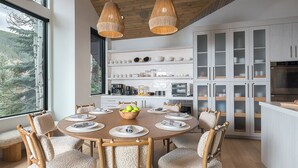 A modern kitchen with white cabinetry, a dining table set for four, and a large window overlooking a forested area. - Sleek, Light-Filled Alpine Escape | Hot Tub, Wine Room | Axiom Retreat (Vail)
