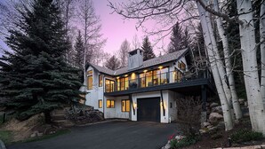 A two-story house with a garage, surrounded by trees and a driveway. - Sleek, Light-Filled Alpine Escape | Hot Tub, Wine Room | Axiom Retreat (Vail)