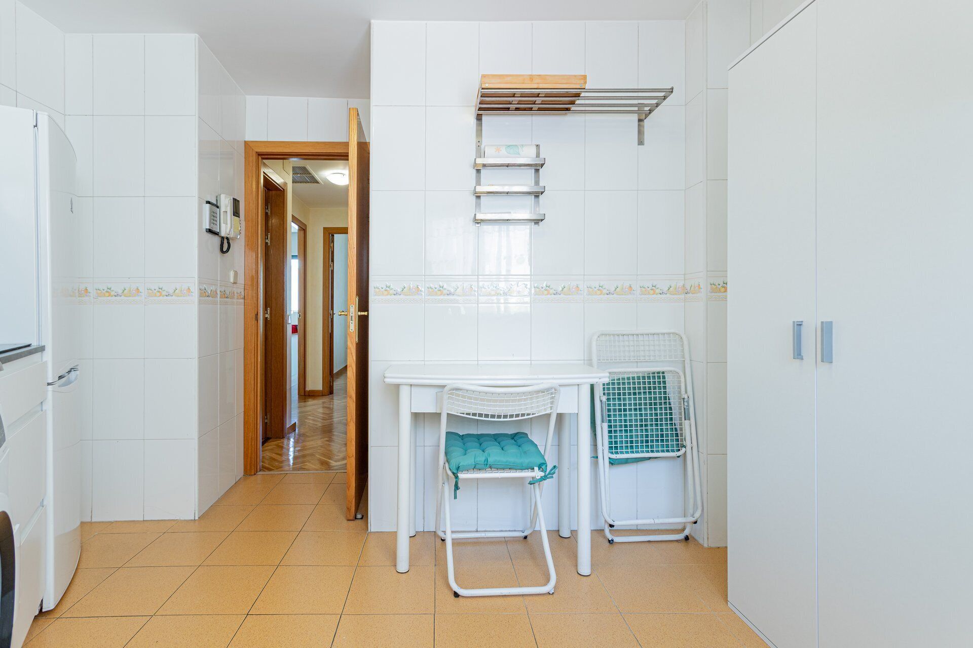 A narrow hallway with a white tiled wall, a wooden door, a white table with a chair, and a drying rack.