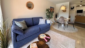 A living room with a blue sofa, a round mirror, a dining area with chairs, and a kitchen area with a TV. - Apartment with Terrace & Pool, Mandelieu (Mandelieu-la-Napoule)