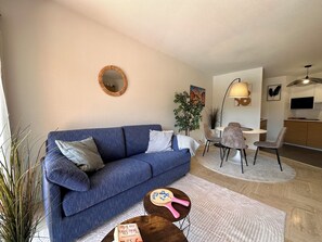 A living room with a blue sofa, a round mirror, a dining area with chairs, and a kitchen area with a TV. - Apartment with Terrace & Pool, Mandelieu (Mandelieu-la-Napoule)
