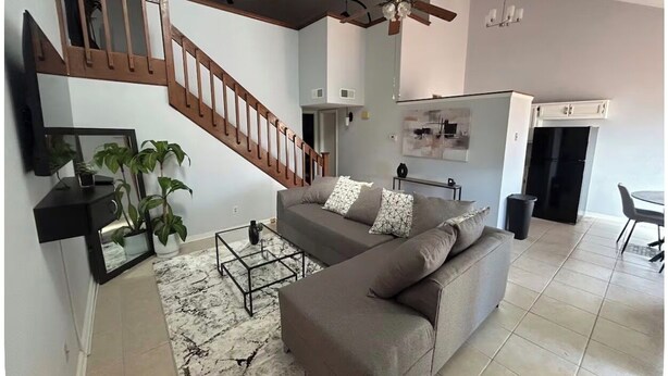 A living room with a sofa, coffee table, and plants. There is a staircase with wooden railings and a ceiling fan.