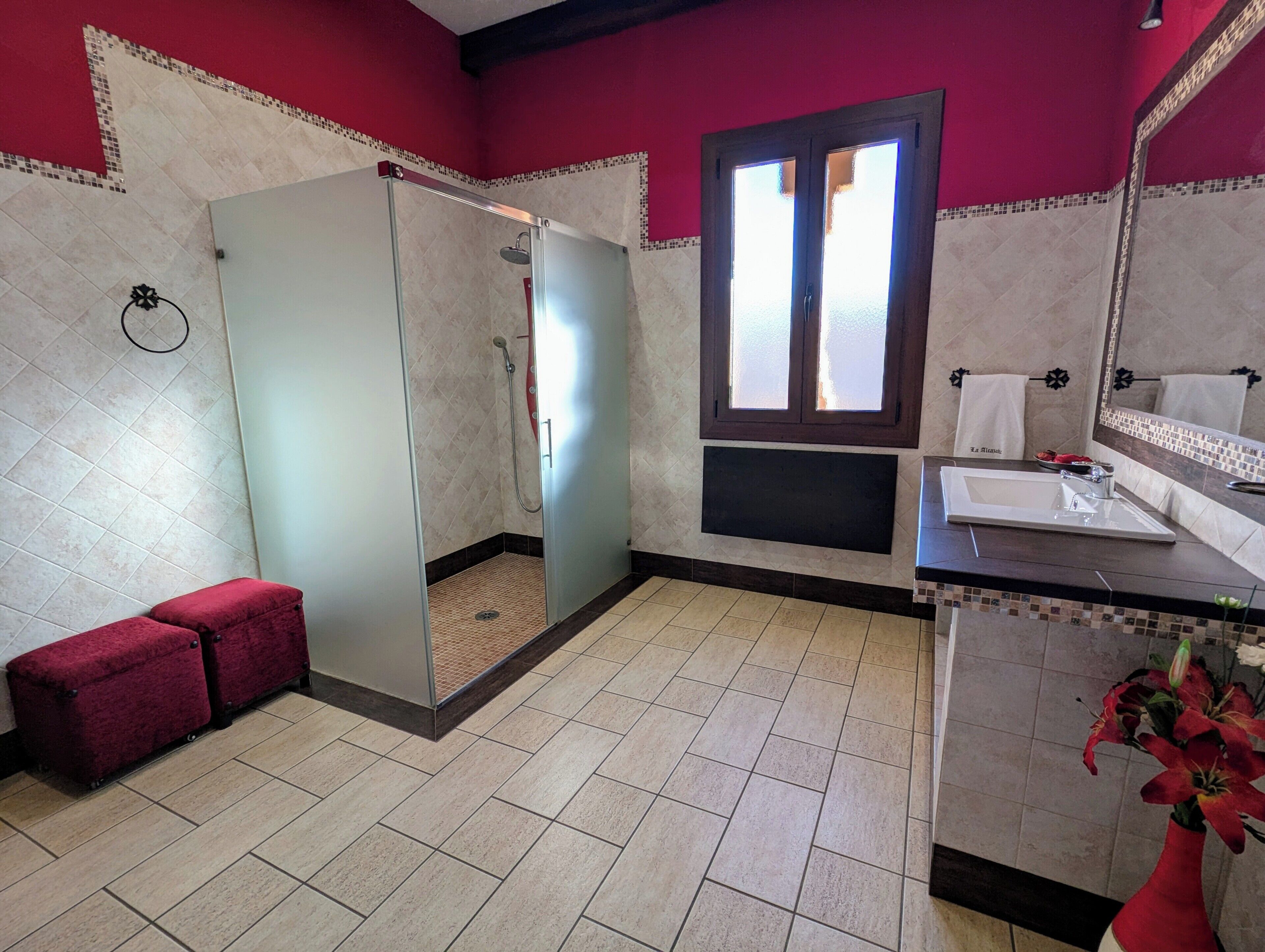 Een badkamer met een douche, wastafel, raam en een rode bank.