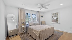 Ein Schlafzimmer mit einem Bett, einem Deckenventilator, einem Fenster mit Vorhängen, einem Nachttisch mit Lampe und einem gerahmten Bild an der Wand.