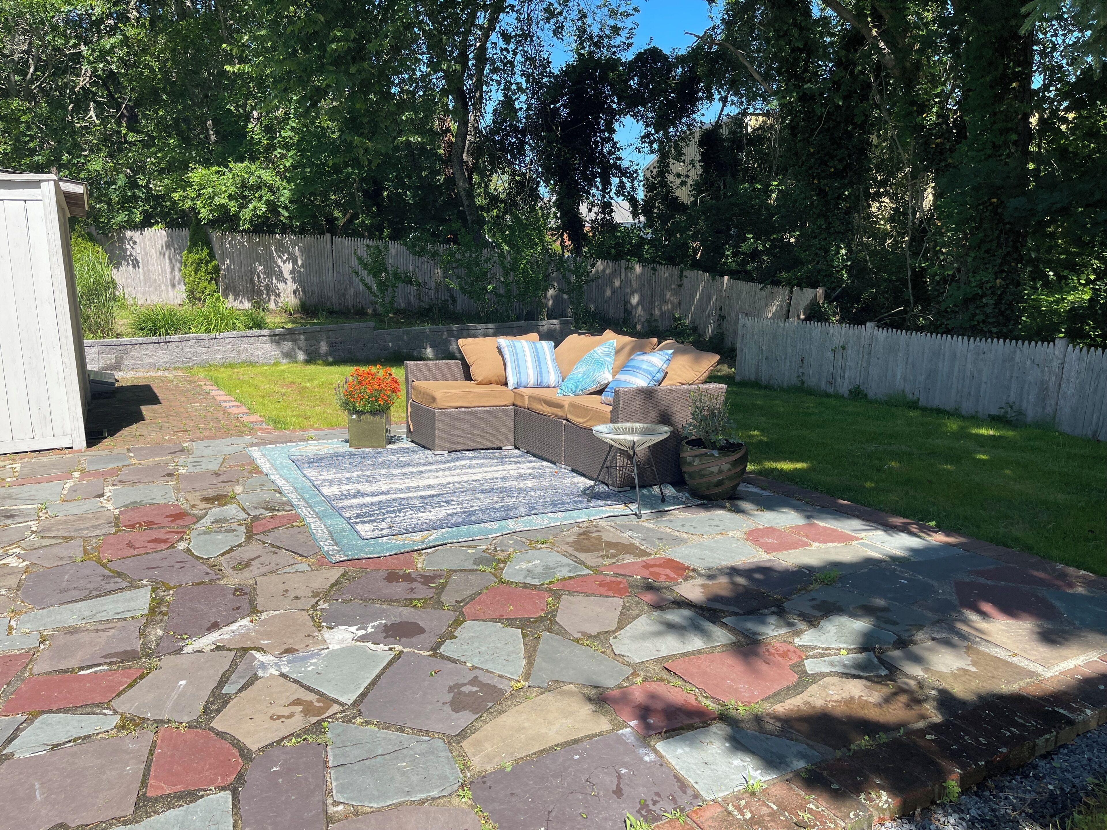 Een patio met een stenen vloer, een rieten bank met kussens, een kleine tafel en een potplant.