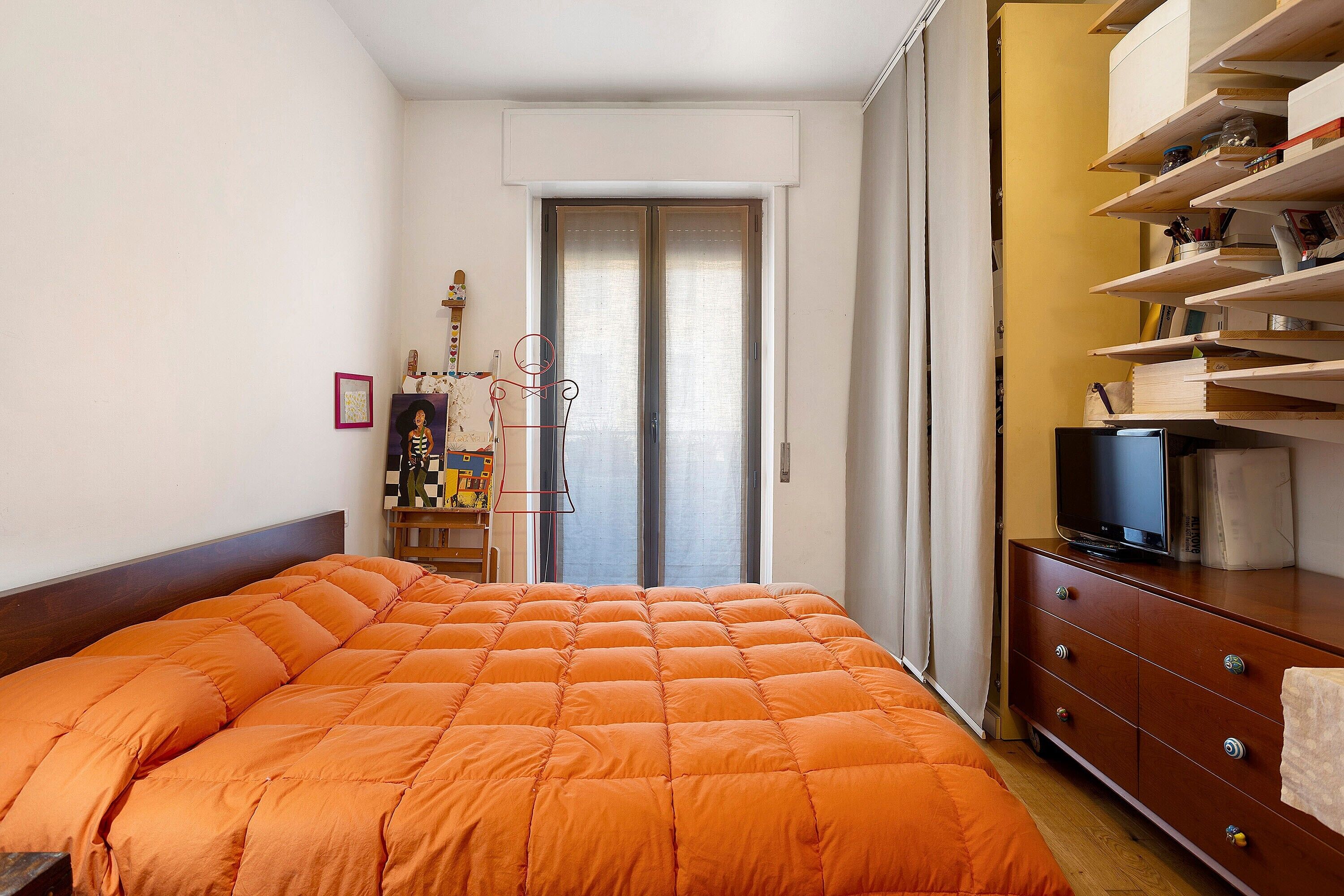 A bedroom with a large orange bed, a wooden dresser, a television, and a door leading to a balcony.