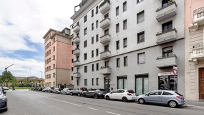 A multi-story building with balconies, parked cars, and a visible storefront with a sign. - Comfortable Apartment Steps from the Red Metro Line (Milano)