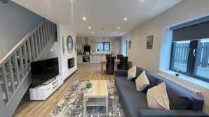A modern living room with a sofa, a coffee table, and a TV. There is a staircase on the left and a kitchen in the background. - Chapel Row 6, Wirksworth Peak District (with pac man machine ) (Wirksworth)