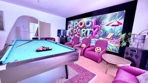 Ein Billardtisch mit Kugeln und Queue in einem Raum, der eine 'POOL PARTY'-Wandmalerei, violette Stühle und einen kleinen Tisch mit einem Lautsprecher zeigt.