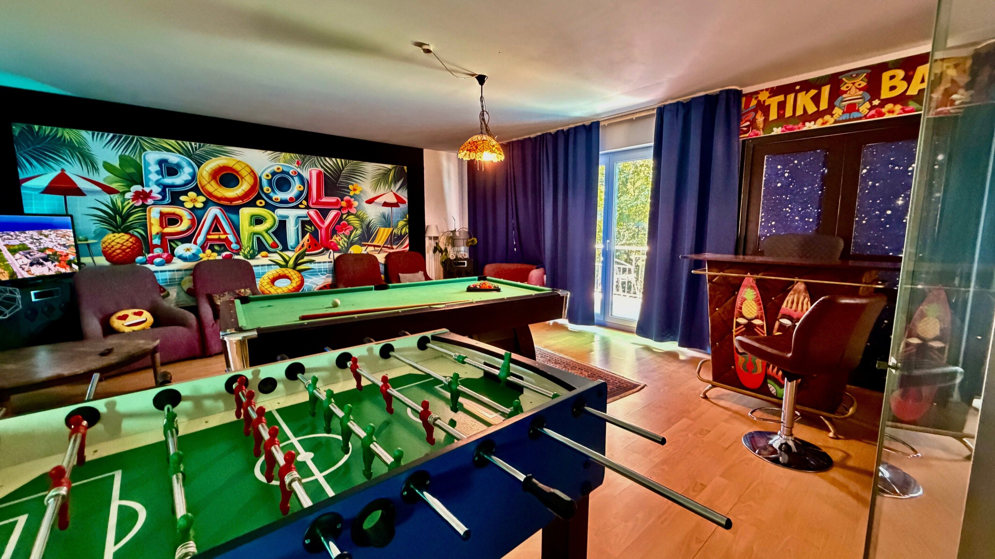 Una habitación con mesa de billar, mesa de futbolín y zona de bar decorada con motivos tiki.