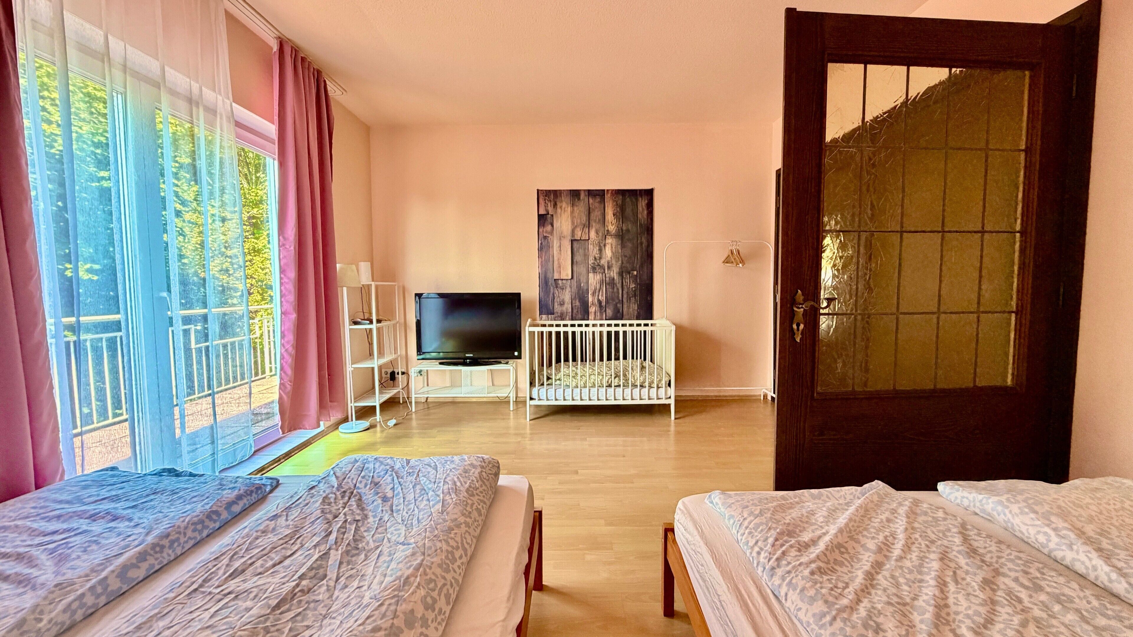 Ein Zimmer mit zwei Betten, einem Fernseher, einem Babybett und einem großen Fenster mit Vorhängen.
