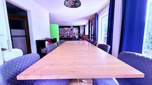 Ein langer Holztisch mit gepolsterten Stühlen in einem Raum mit großen Fenstern und einer farbenfrohen Wandmalerei.