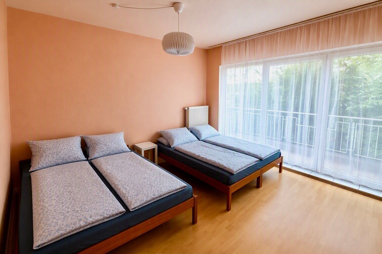 Ein Zimmer mit zwei Betten, einem Heizkörper, einem Fenster mit durchsichtigen Vorhängen und einer Pendelleuchte.