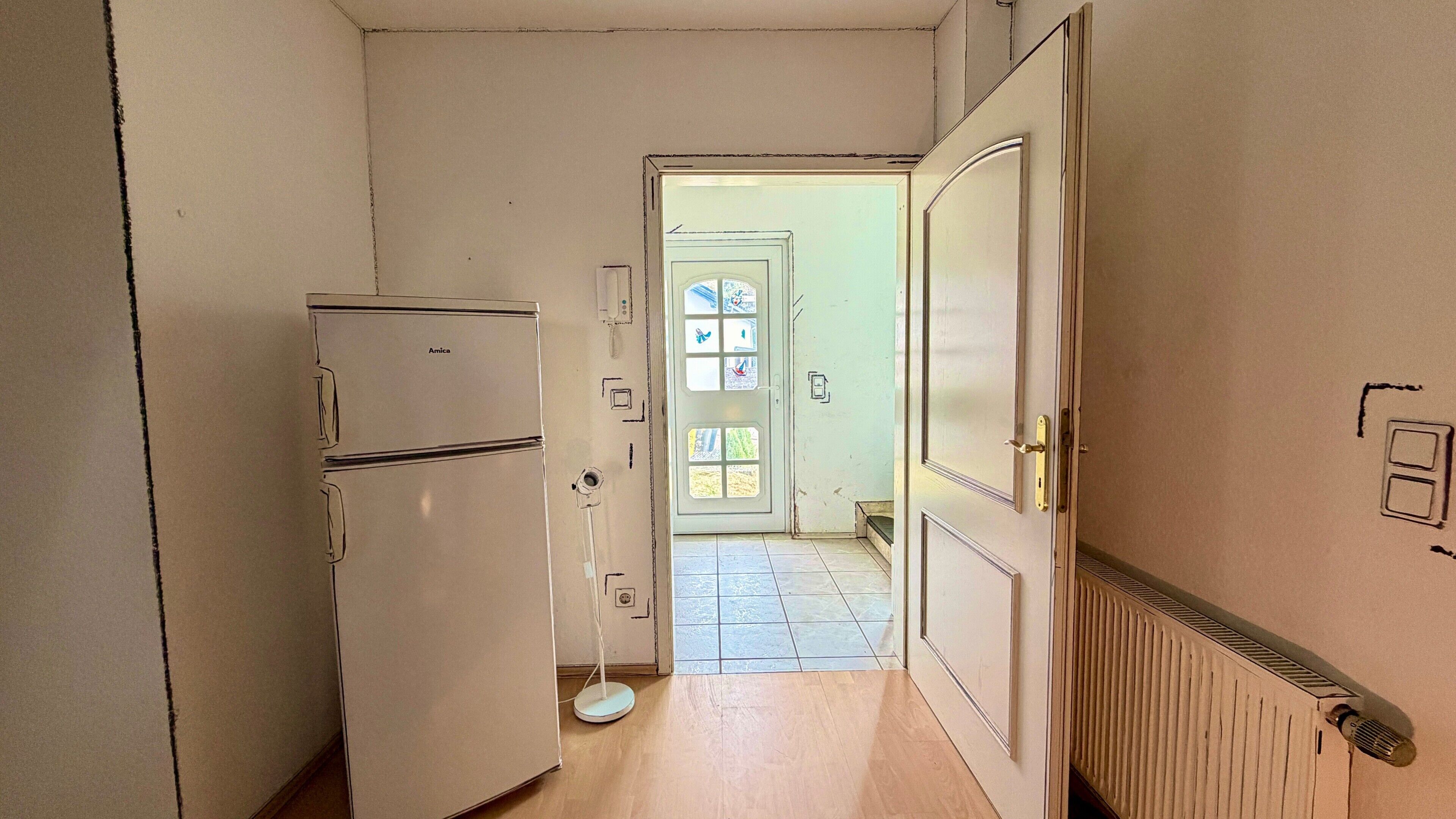 Ein schmaler Flur mit einem weißen Kühlschrank, einer Tür zu einem Raum mit Fliesenboden und einem Heizkörper an der Wand.