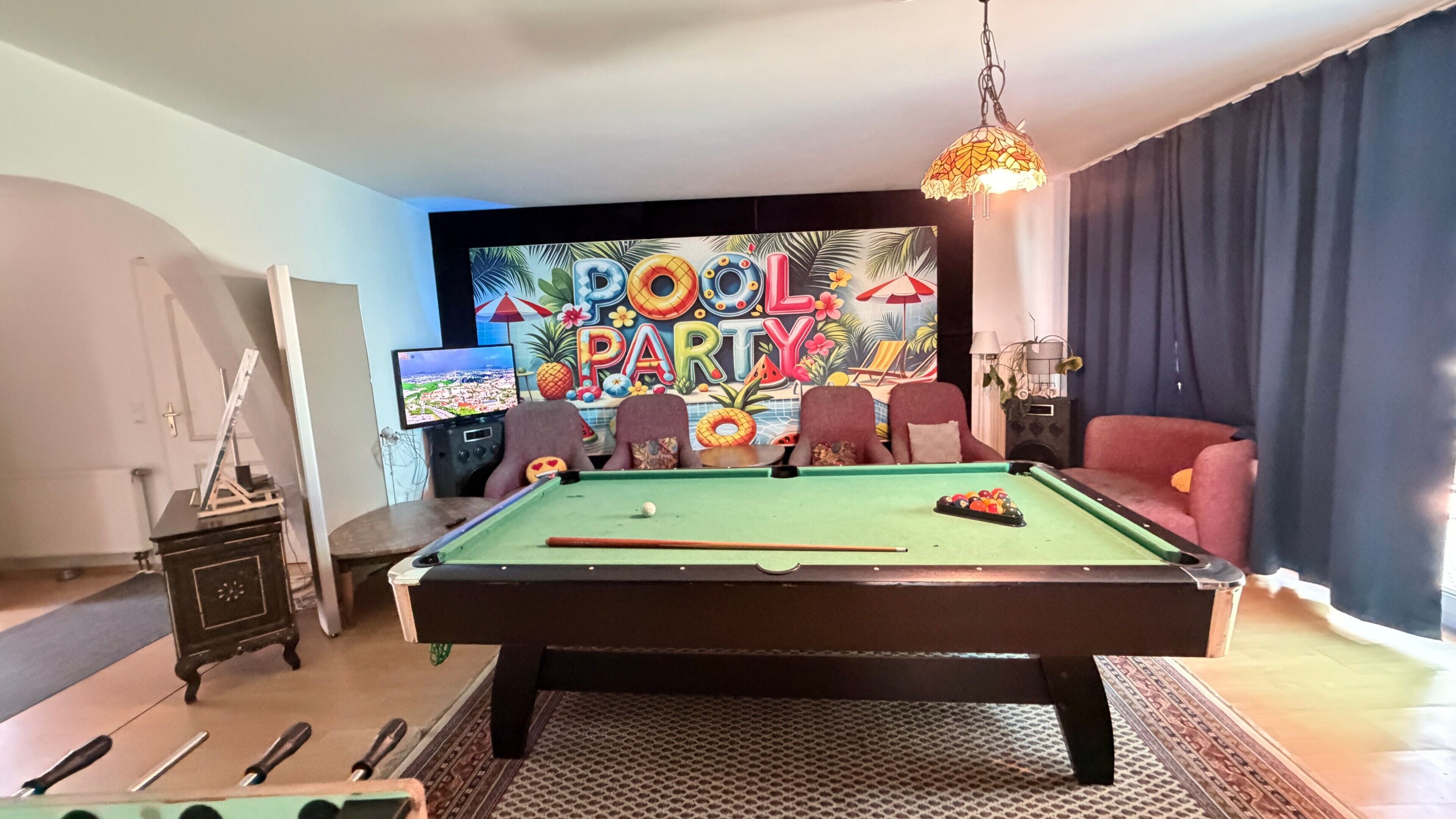 A pool table with a pool cue, a billiards rack, and a cue ball. There is a television displaying a cityscape. A pool table with a colorful backdrop and the words 'POOL PARTY' is visible.