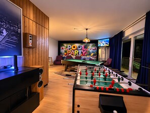 A room with a pool table, foosball table, a television, and a large screen.