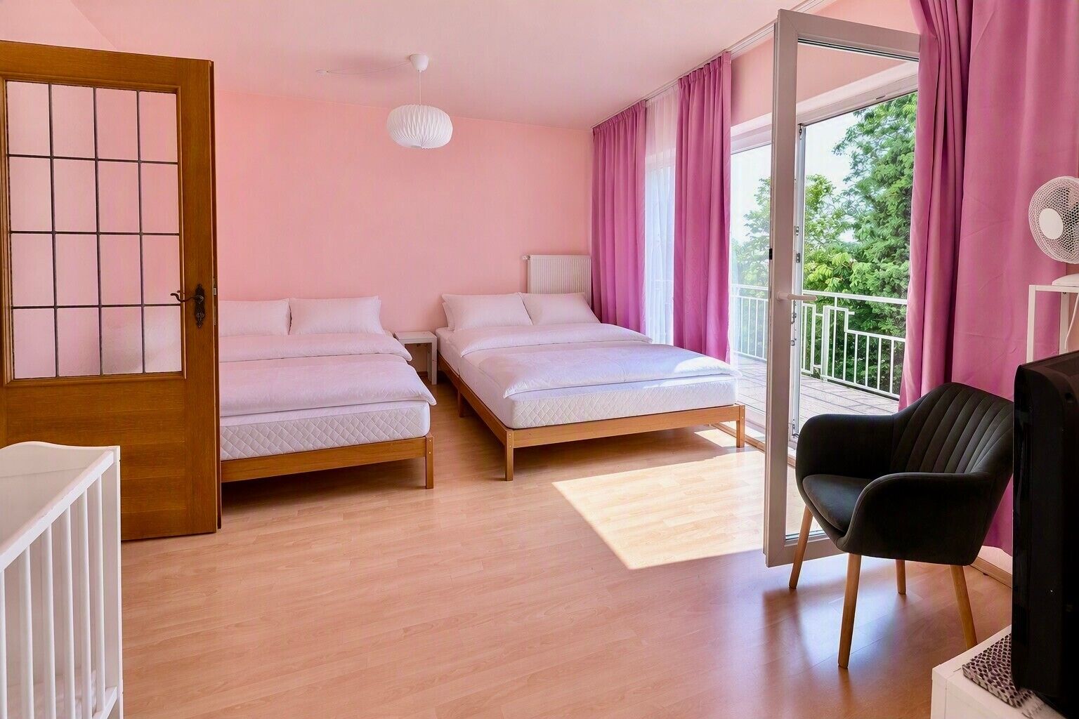 Ein Schlafzimmer mit zwei Betten, einer Holztür, einem weißen Deckenventilator und einem Balkon mit rosafarbenen Vorhängen.