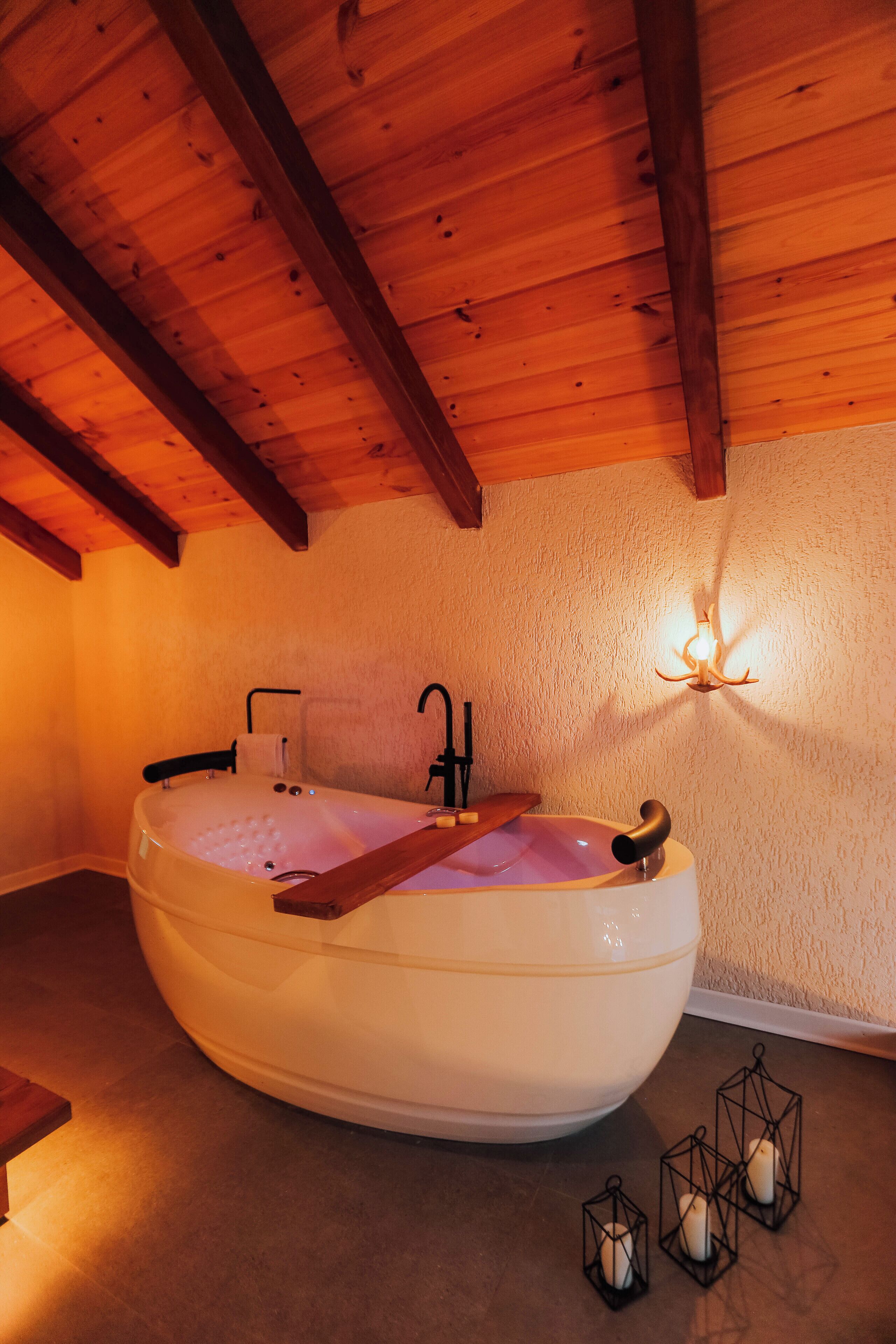 A room with a large bathtub, wooden beams on the ceiling, a wall-mounted light fixture, and candles on the floor.