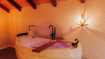 A room with a large bathtub, wooden beams on the ceiling, a wall-mounted light fixture, and candles on the floor.