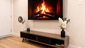 Ein Zimmer mit an der Wand montiertem Fernseher, der ein Bild eines Kamins zeigt, einem Holzkonsolentisch mit Blumenvase und einem Fußventilator.