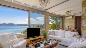 A spacious living room with a large window offering a view of the sea, a white sofa, a wooden coffee table, and a television.