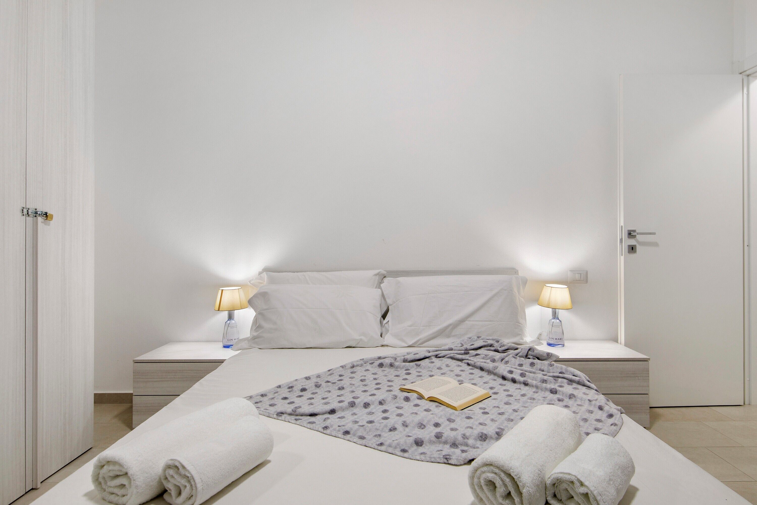 Un lit bien fait, avec une couverture, des oreillers et un livre, deux lampes de chevet et une porte blanche.