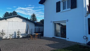 Kaksikerroksinen valkoinen rakennus, jossa on katettu terassi, aidattu piha ja kirkas sininen taivas.