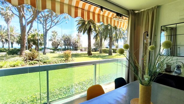 A room with a large window, a table with a vase of plants, and a view of a well-maintained outdoor area with palm trees and a fence. - REF DOLOS BY PALMES D OR PROPERTIES (Cannes)