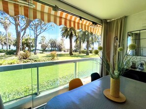 A room with a large window, a table with a vase of plants, and a view of a well-maintained outdoor area with palm trees and a fence. - REF DOLOS BY PALMES D OR PROPERTIES (Cannes)