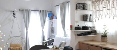 A dining area with a table set for two, a hanging light fixture, and a kitchen area with shelves.