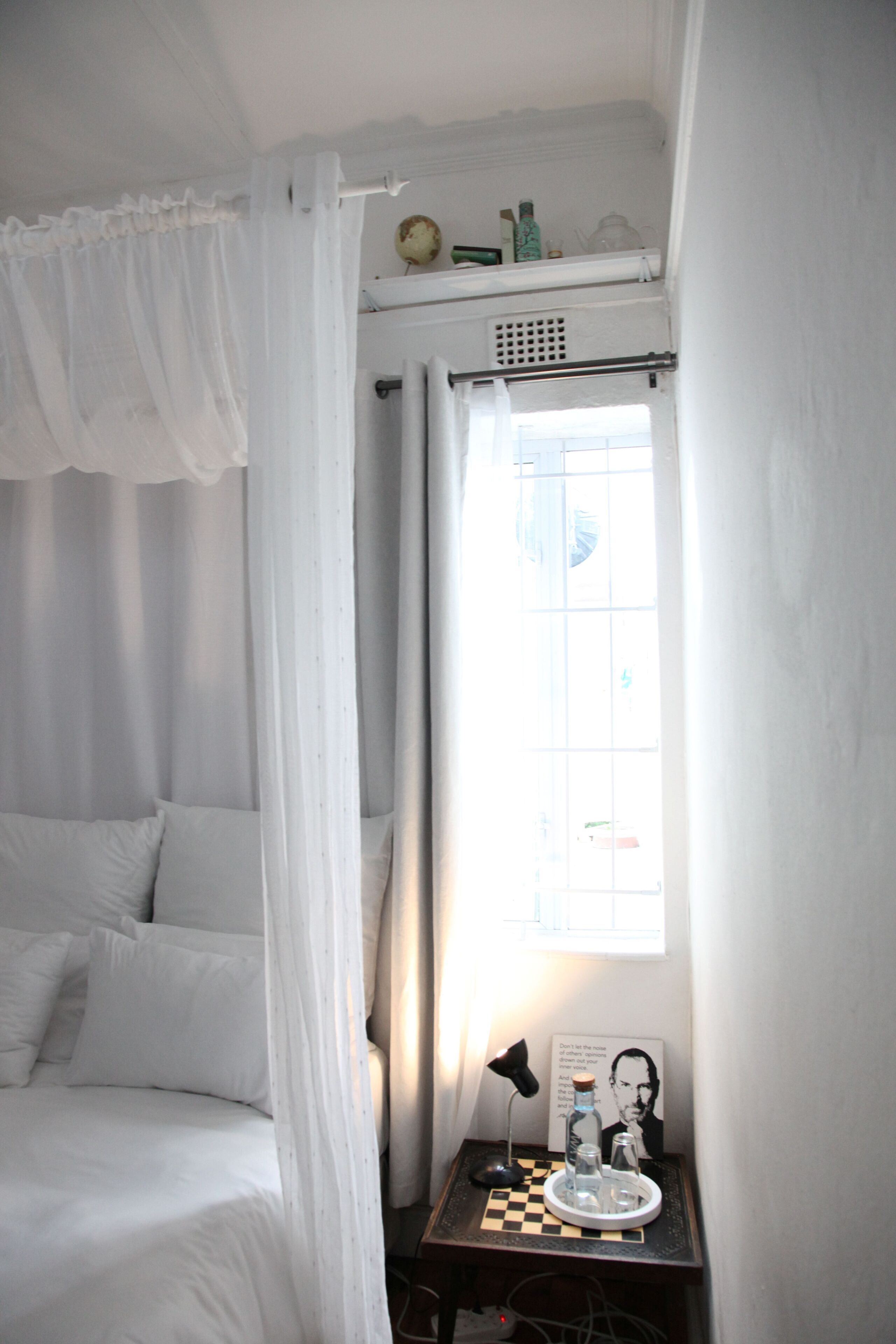 Un dormitorio con cama, mesita de noche con lámpara, ventana con cortinas y un estante con objetos.