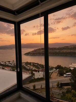 Ein Zimmer mit großem Fenster und Blick auf eine Küstenlandschaft bei Sonnenuntergang.