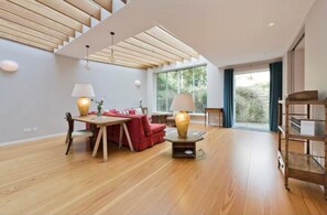 Ein geräumiges Zimmer mit Holzboden, einem Essbereich mit Tisch und Stühlen, einem roten Sofa und einem großen Fenster mit Blick auf Grünflächen.