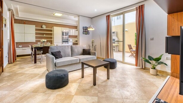 A modern living room with a sofa, ottoman, and coffee table, a dining area with chairs, a kitchen with cabinets and appliances, and a sliding glass door leading to an outdoor area.