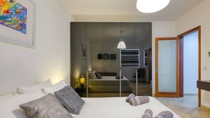A modern hotel room with a large bed, a mirror, a sofa, and a view of the adjacent room.