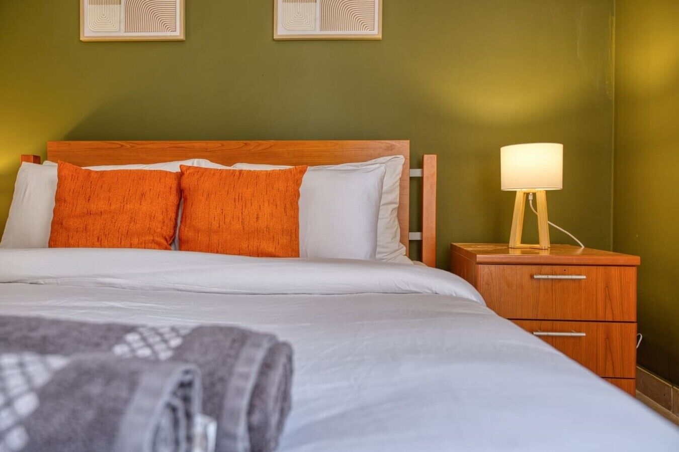 A neatly made bed with white and orange pillows, a wooden nightstand with a lamp, and two framed artworks on the wall.