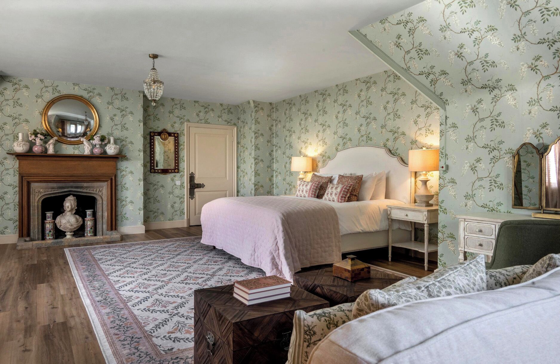 Un dormitorio con cama, lámparas de noche, chimenea, espejo y papel tapiz floral.
