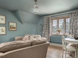 A room with a beige sofa, a dining table with chairs, a window with patterned curtains, and framed wall art. - Whistler Rooms (Peterborough)