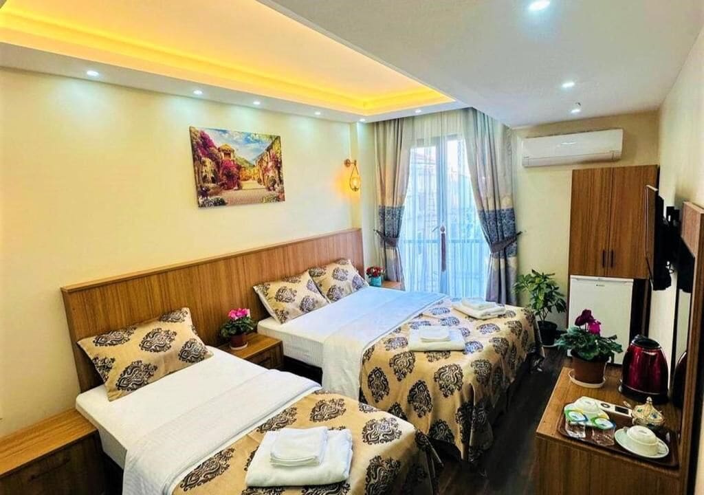 Habitación de hotel con dos camas, una mesita con hervidor, una planta en maceta y un cuadro en la pared.