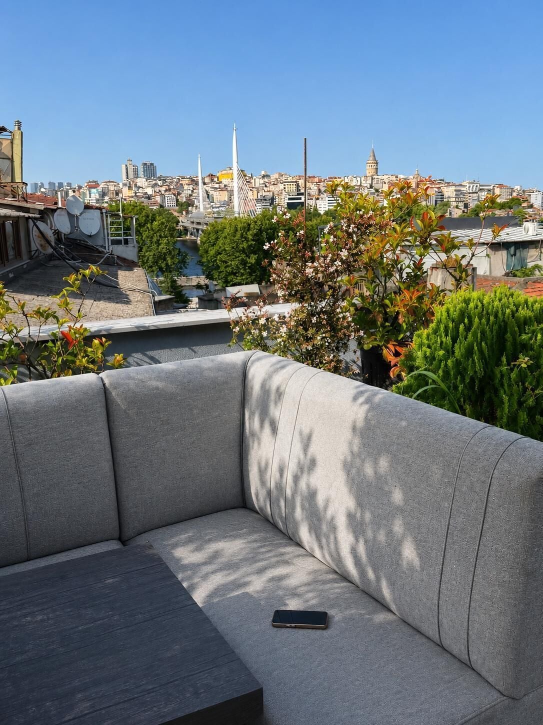 A rooftop terrace with a grey sofa, a smartphone on the seat, and a view of the cityscape.